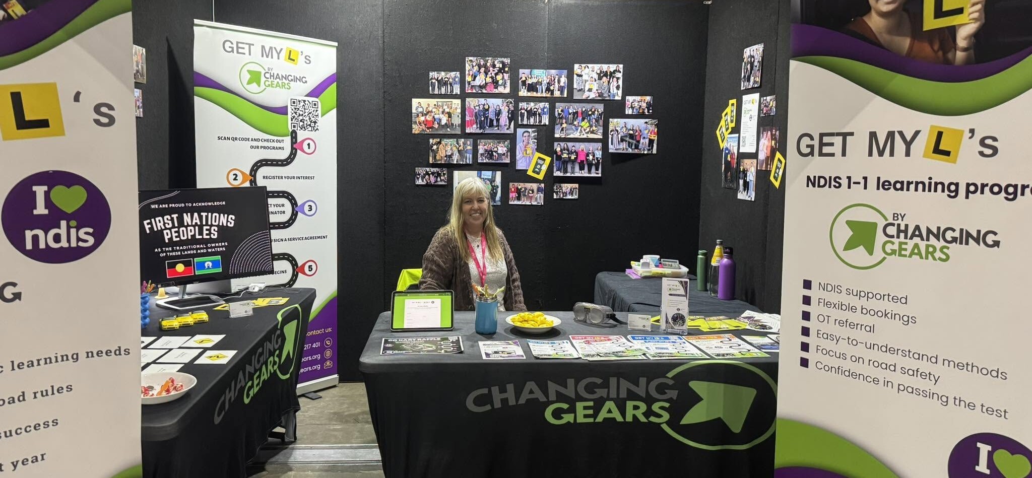 Linda smiling behind the Changing Gears table at last year's expo, with Get My L's banners on each side and a wall of photos of past participants holding their L plates.